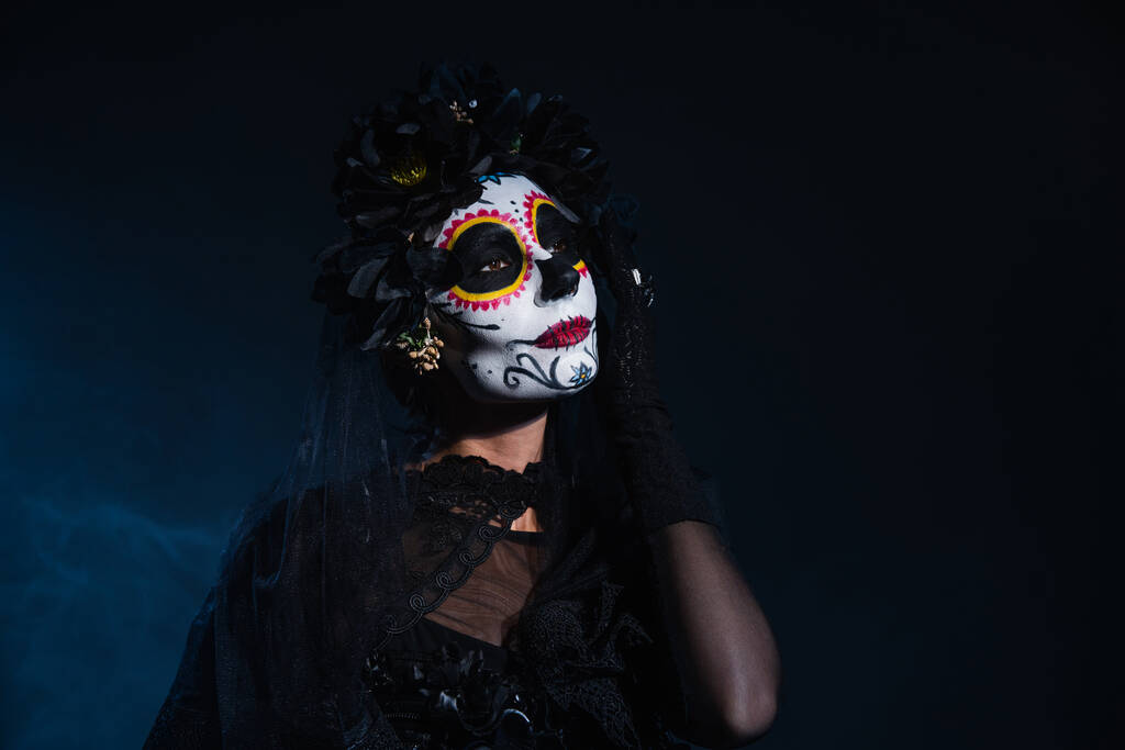 Frau mit Totenkopf-Halloween-Make-up hält Hand in Gesichtsnähe auf schwarzem und blauem Hintergrund - Foto, Bild