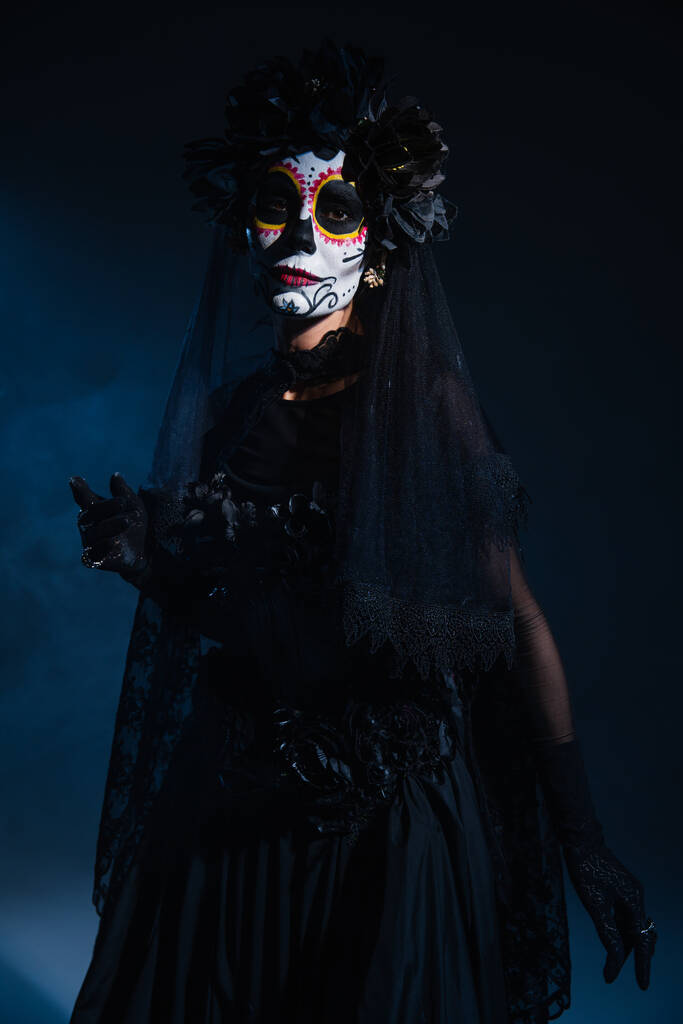 mujer en maquillaje de santa muerte y traje espeluznante con velo de encaje negro sobre fondo azul oscuro - Foto, Imagen