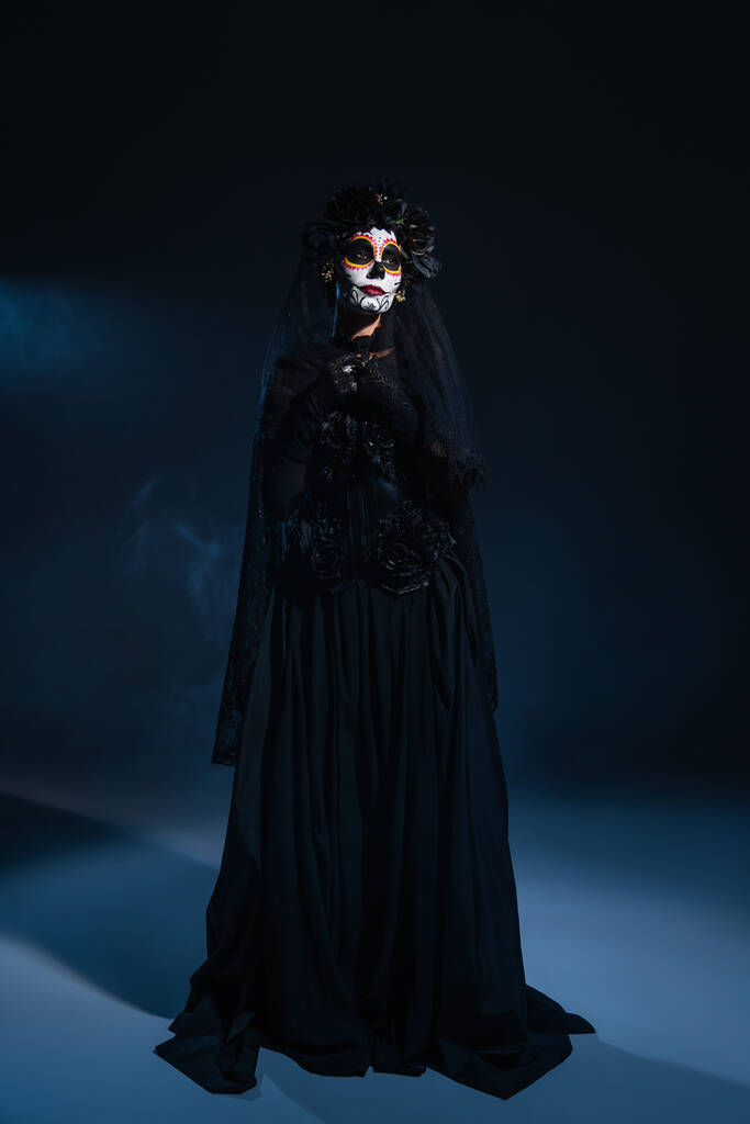Siyah cadı elbisesi ve cadılar bayramı makyajı giymiş koyu renk arka planda mavi ışıklı bir kadın. - Fotoğraf, Görsel