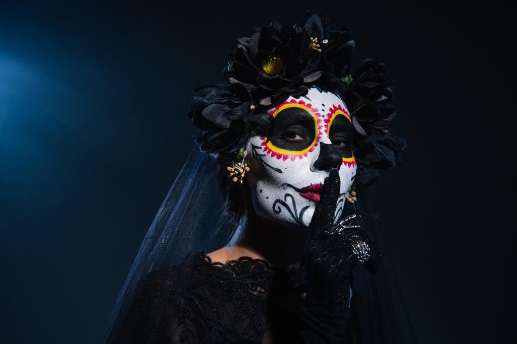 mujer en espeluznante maquillaje de halloween y corona negra con velo mostrando signo de silencio sobre fondo azul oscuro - Foto, imagen
