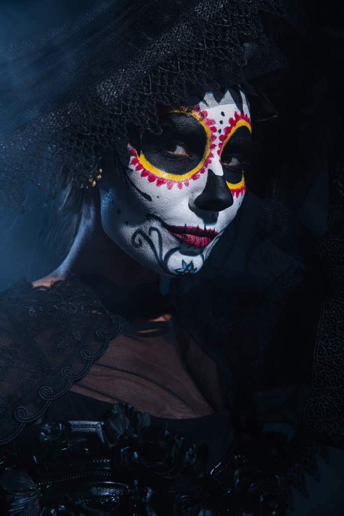 vrouw in traditionele dag van de dood make-up en kostuum kijken naar camera op donkerblauwe achtergrond - Foto, afbeelding