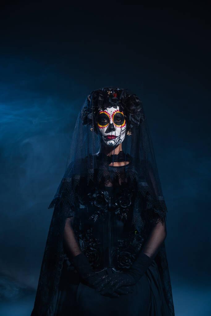 вид спереду жінки в чорному костюмі відьми і страшний макіяж на Хелловін на темному тлі з блакитним туманом
 - Фото, зображення