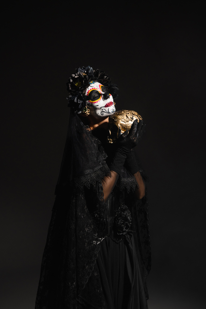 Frau mit Zuckerschädel-Make-up trägt dunkles Kostüm mit Schleier und hält goldenen Totenkopf isoliert auf schwarz - Foto, Bild