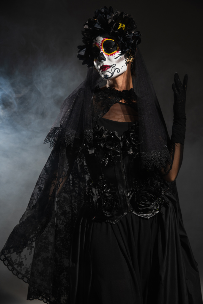 vrouw in zwart heks kostuum en griezelige halloween make-up poseren op donkere achtergrond met mist - Foto, afbeelding