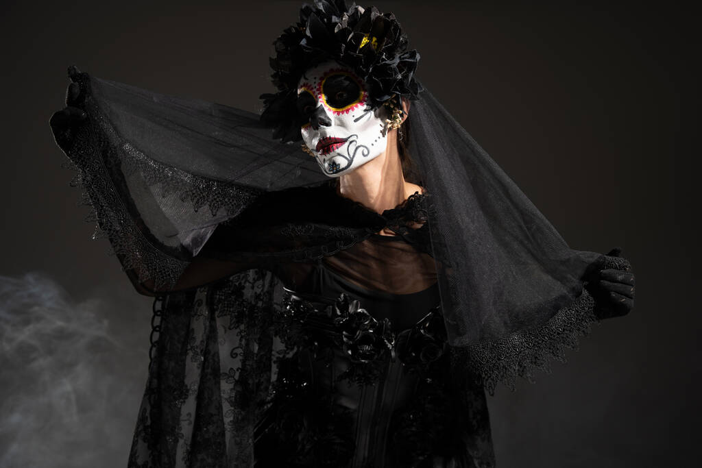 женщина в жутком Хэллоуин макияж и костюм ведьмы держа черный кружевной вуаль на темном фоне с дымом - Фото, изображение