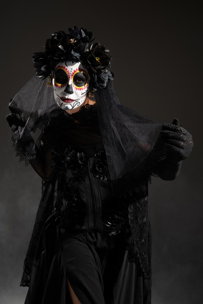 Cadılar bayramı şekerli kafatası makyajlı ve siyah kostümlü bir kadın karanlık arka planda kameraya bakıyor. - Fotoğraf, Görsel