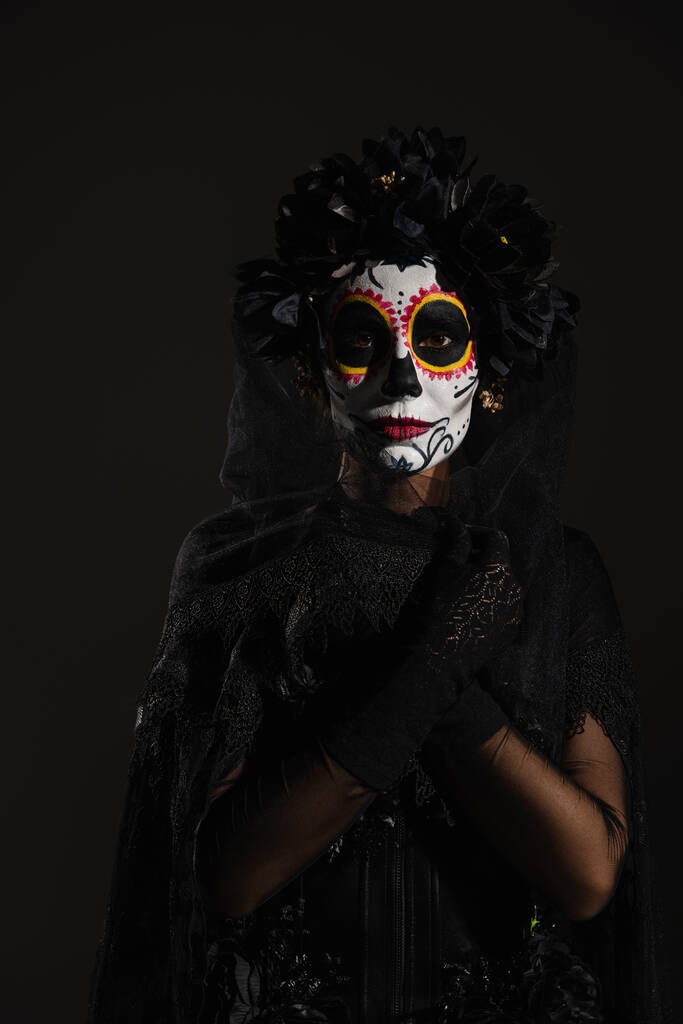 Frau mit mexikanischem Tag des toten Make-ups hüllt sich in dunklen Schleier und blickt vereinzelt in die Kamera auf schwarz - Foto, Bild