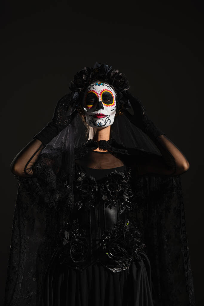 kobieta w makijażu Catrina i Santa Muerte kostium dotykając ciemny wieniec z welonem izolowane na czarno - Zdjęcie, obraz