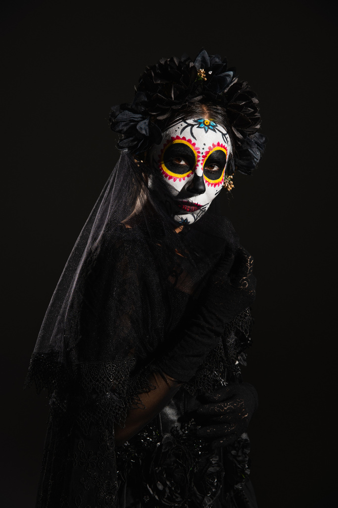donna in tradizionale trucco Santa Muerte e costume scuro con ghirlanda guardando la fotocamera isolata su nero - Foto, immagini
