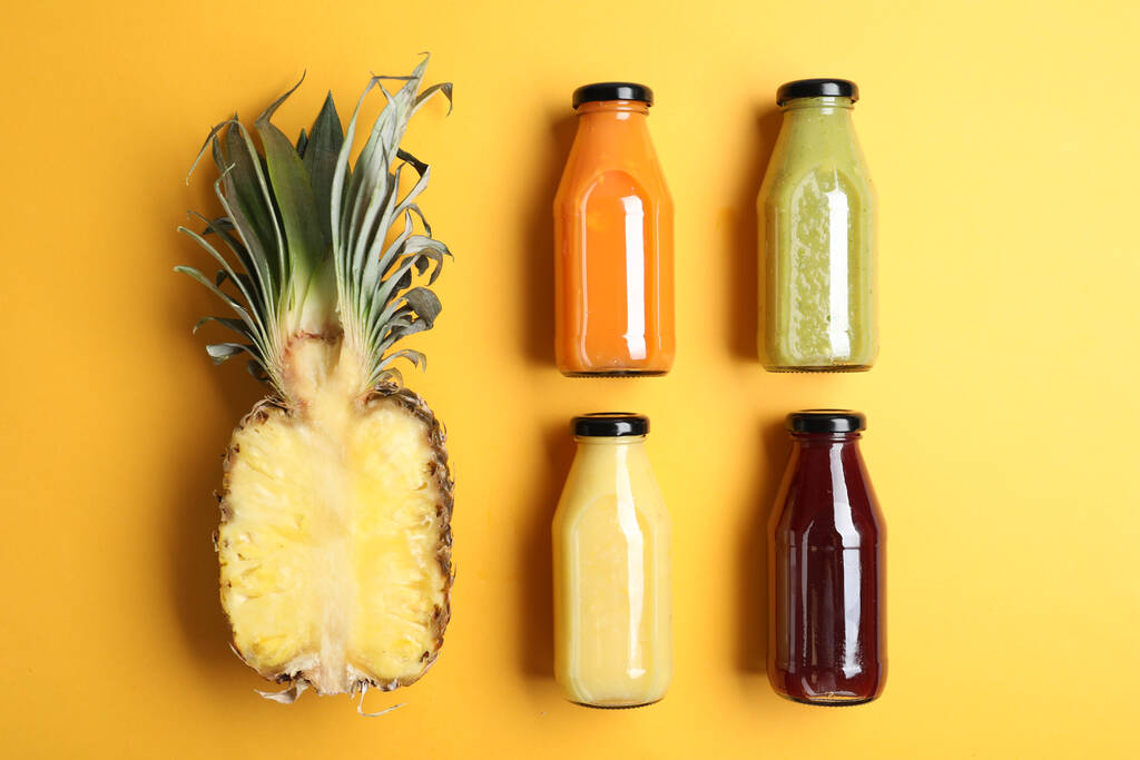 Bottles with delicious colorful juices and cut pineapple on yellow background, flat lay - Foto, Bild