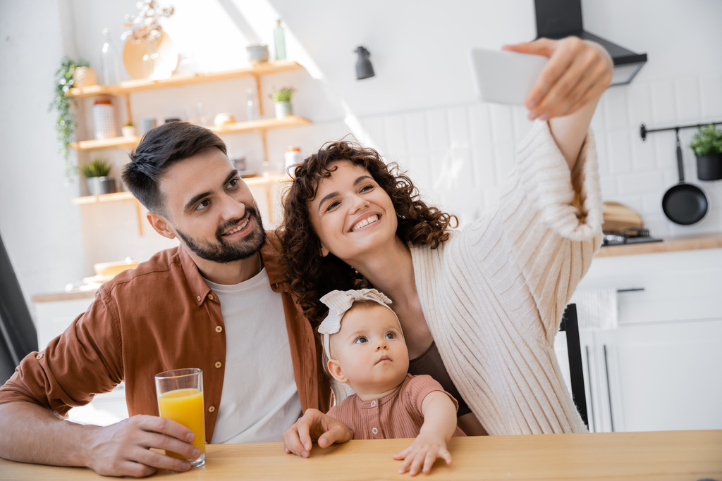 σγουρή γυναίκα που βγάζει selfie με ευτυχισμένο σύζυγο και νήπιο κόρη στο σπίτι - Φωτογραφία, εικόνα
