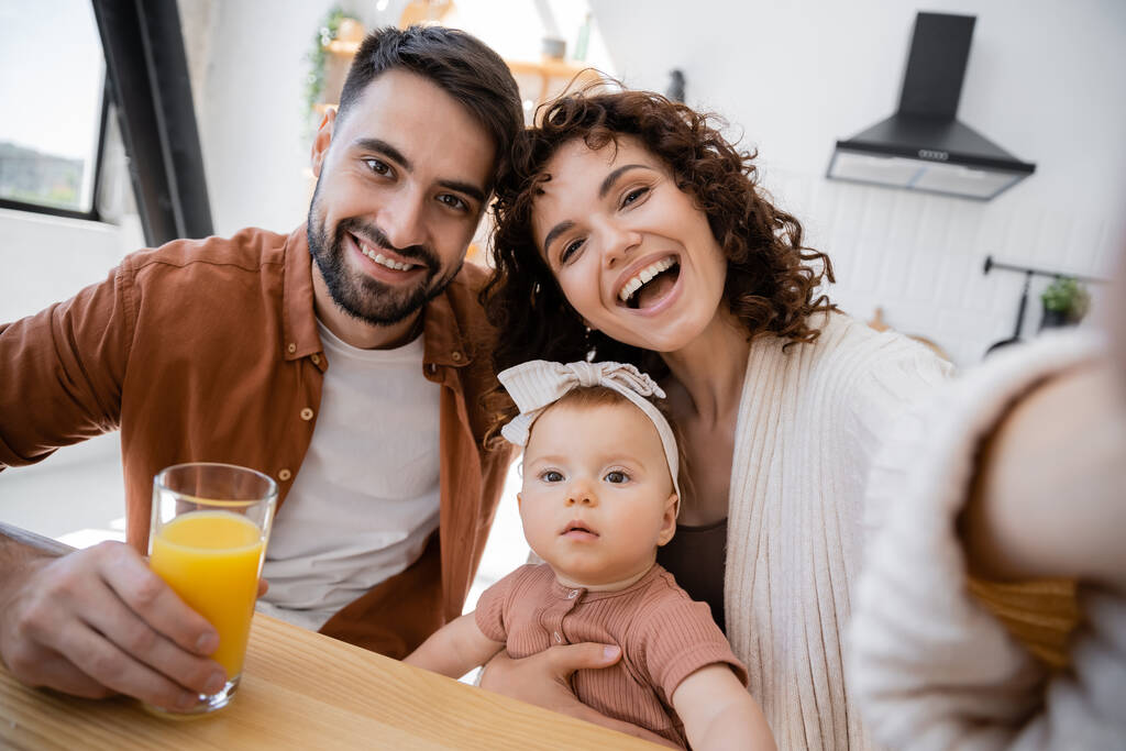 curly woman smiling near cheerful husband and infant daughter while looking at camera  - Photo, Image