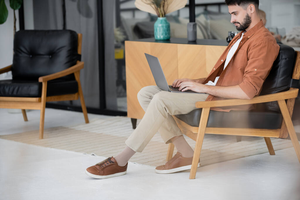 full length of bearded freelancer sitting on leather armchair and using laptop  - Photo, Image
