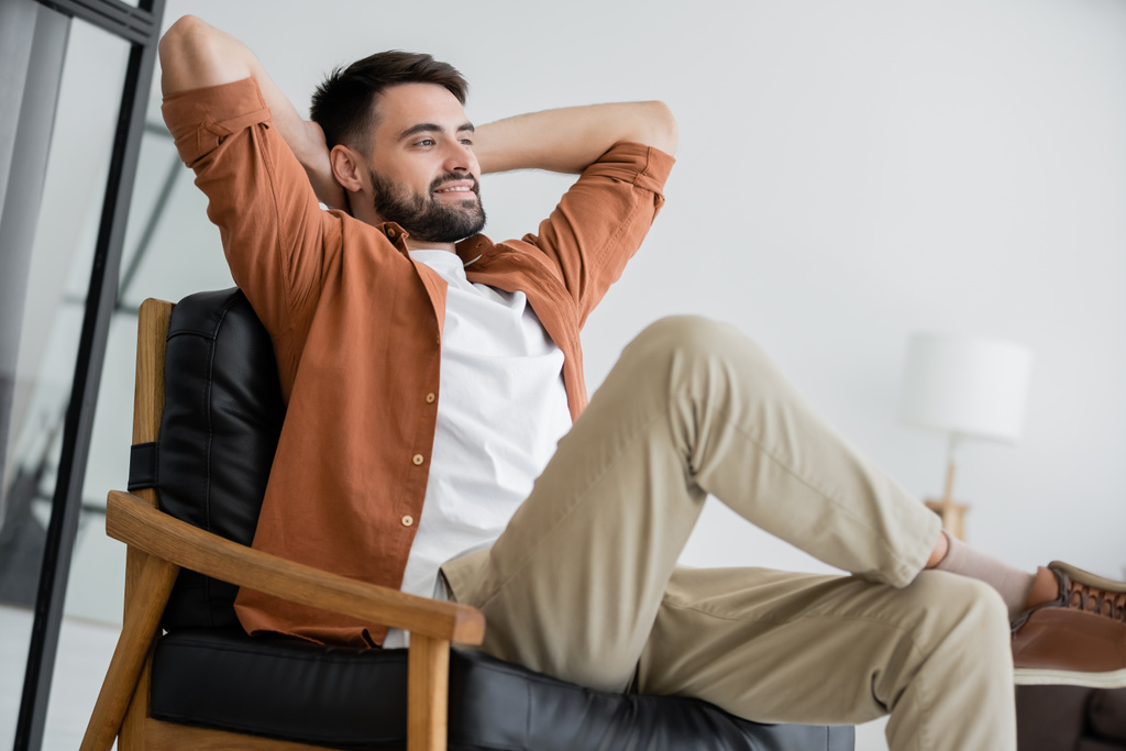 happy and bearded man sitting on comfortable leather armchair in living room  - Photo, Image