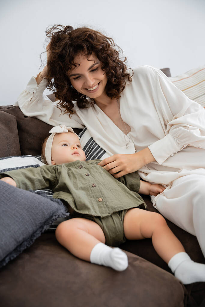 felice madre in abito sorridente vicino figlia neonata in fascia seduta sul divano - Foto, immagini