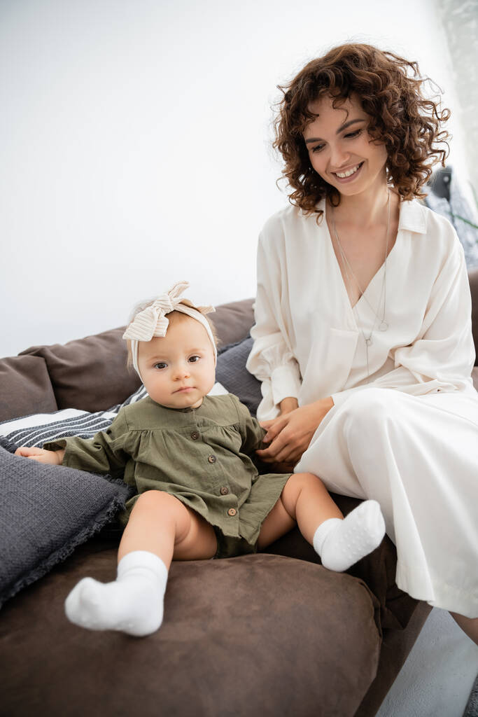 alegre madre en vestido sonriendo cerca de hija bebé en diadema sentado en sofá - Foto, Imagen