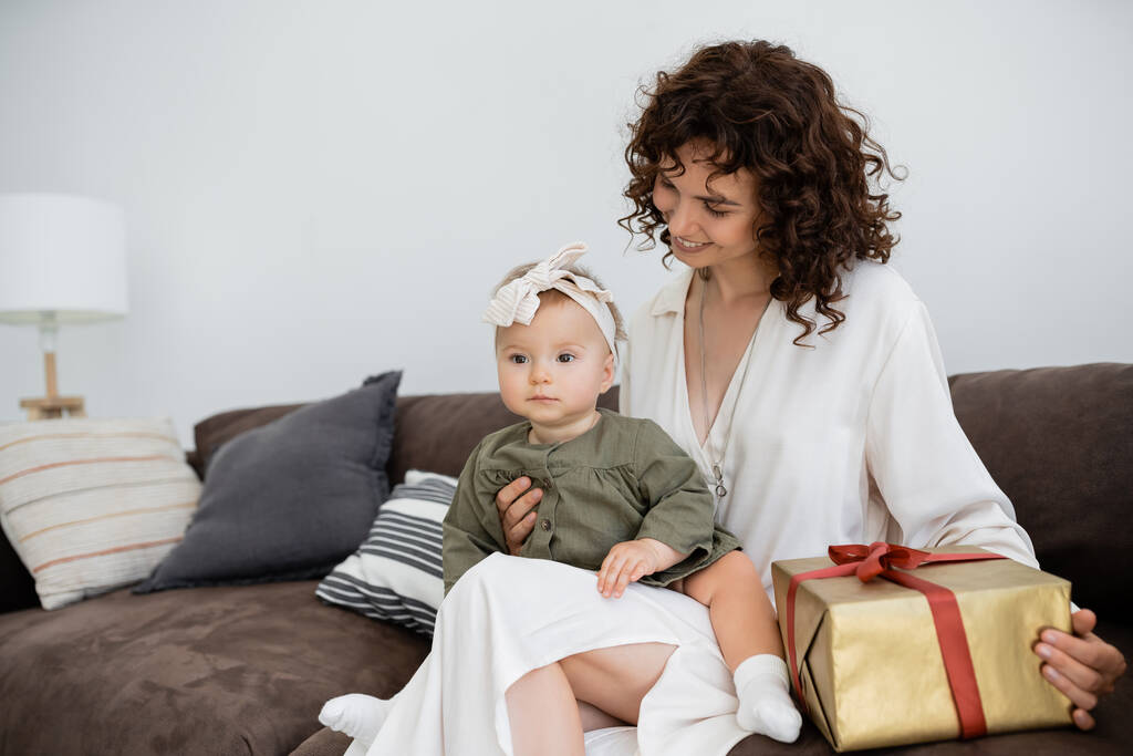 Neşeli ve kıvırcık elbiseli anne saç bandı takmış küçük kızının yanında hediye paketi taşıyor. - Fotoğraf, Görsel