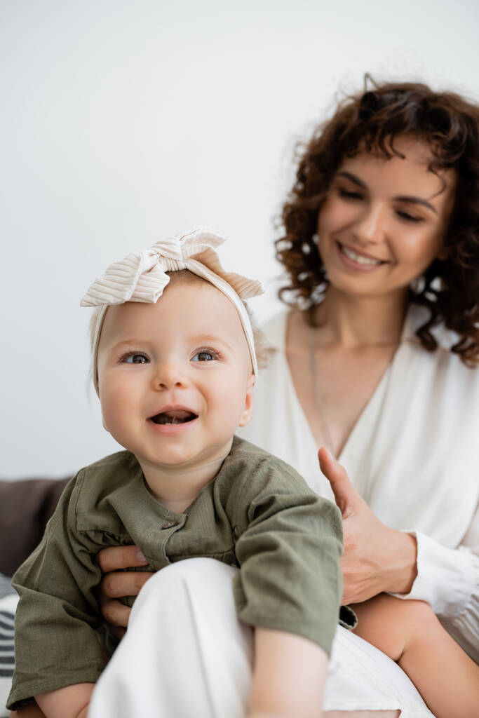 счастливая мама с вьющимися волосами улыбается, держа на руках малышку в повязке на голове  - Фото, изображение