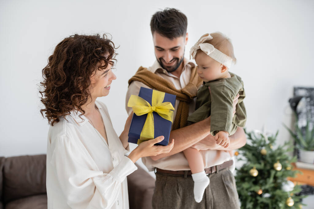 glückliche Mutter hält Geschenkbox in der Nähe fröhlicher Ehemann hält kleine Tochter an Weihnachten  - Foto, Bild