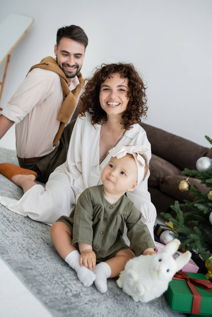 padres felices y linda niña sentada con juguete suave cerca del árbol de Navidad decorado con regalos  - Foto, Imagen