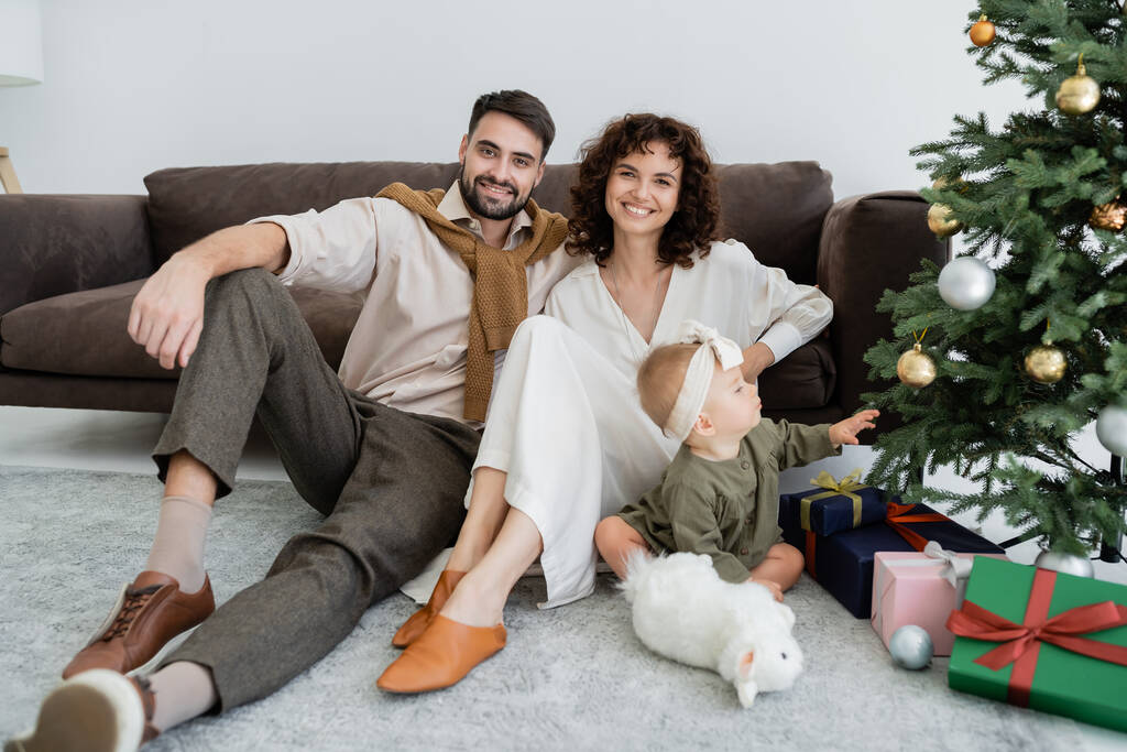 happy parents sitting near infant girl and decorated christmas tree with presents  - Photo, Image