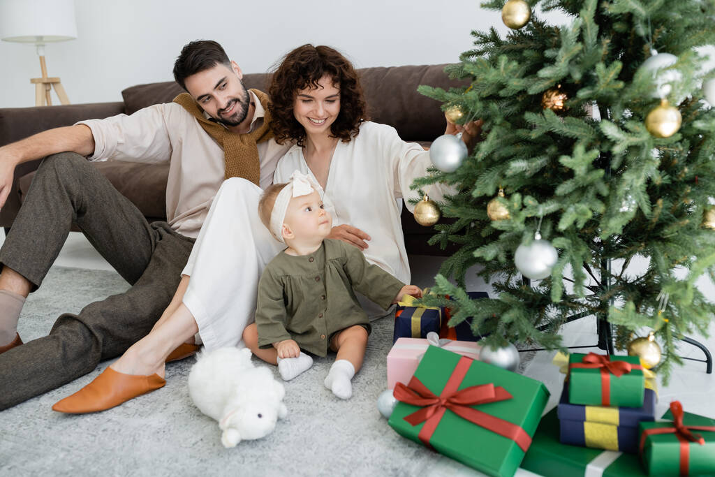 genitori felici e bambina seduta vicino a scatole regalo sotto l'albero di Natale decorato  - Foto, immagini