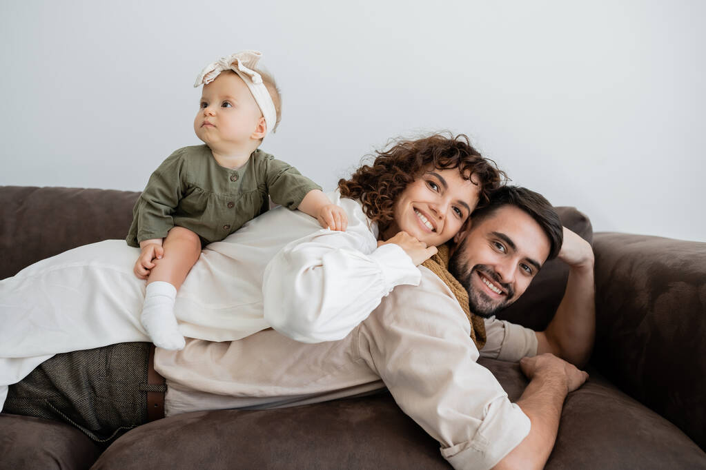 veselá a kudrnatá žena a kojenecká dívka ležící na zádech vousatého manžela v obývacím pokoji - Fotografie, Obrázek