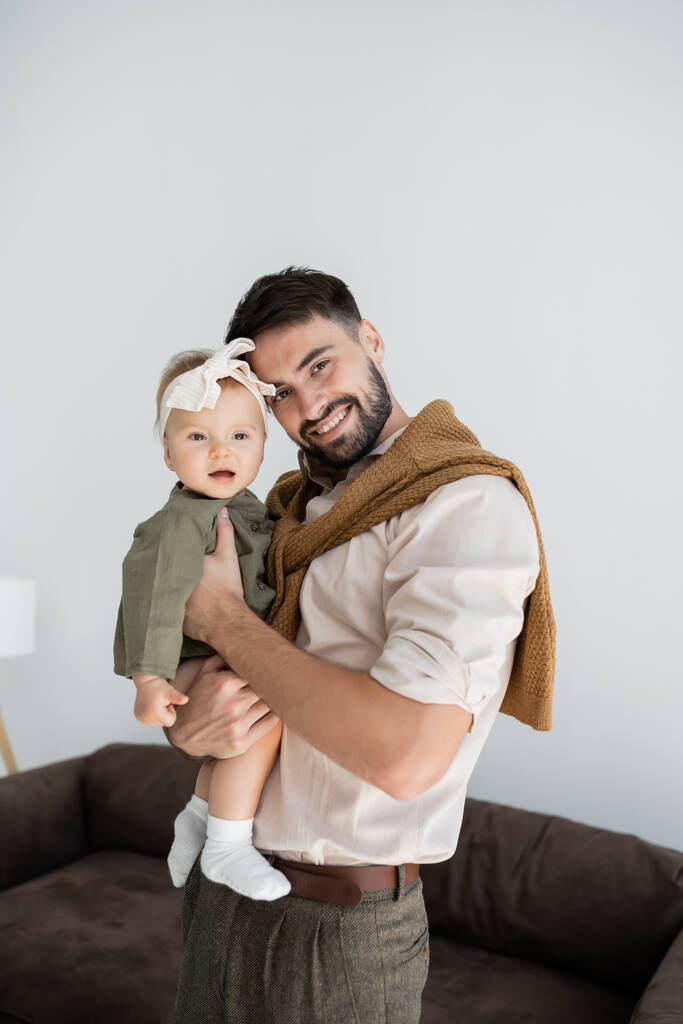 θετική γενειοφόρος άνδρας εκμετάλλευση στην αγκαλιά κόρη του μωρού σε φόρεμα και κεφαλόδεσμο   - Φωτογραφία, εικόνα