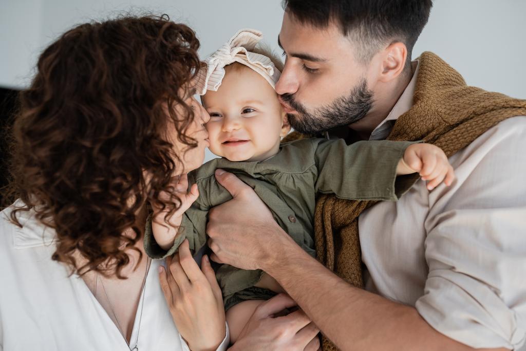 uomo barbuto e donna riccia baciare guance di figlia felice bambino in fascia  - Foto, immagini