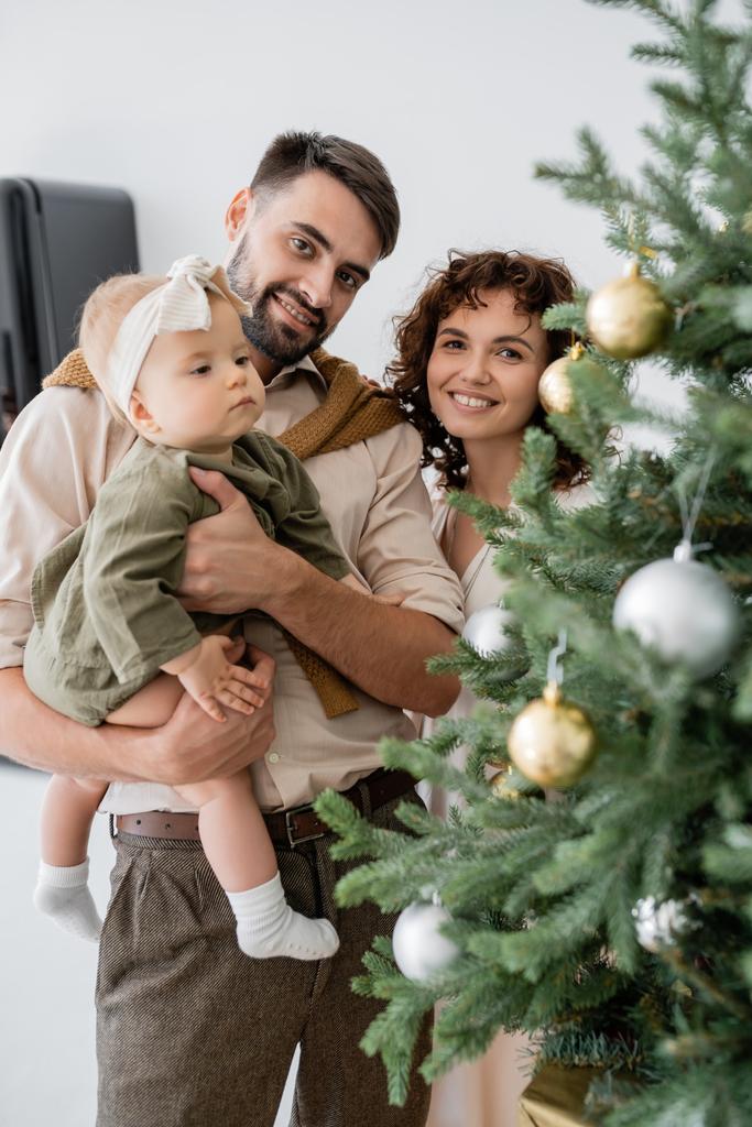 felice padre che tiene figlia neonato vicino alla moglie allegra e decorato albero di Natale  - Foto, immagini