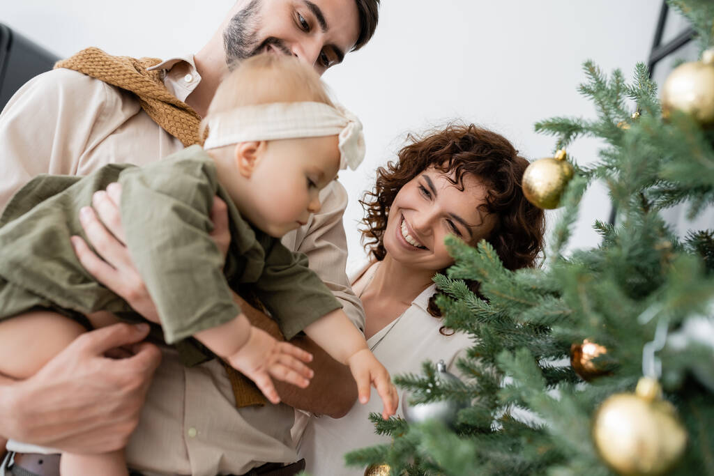glücklicher Vater hält kleine Tochter neben lächelnder Frau und geschmücktem Weihnachtsbaum  - Foto, Bild