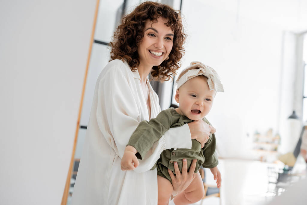 donna felice e riccia in abito bianco che tiene in braccio la figlia neonata - Foto, immagini