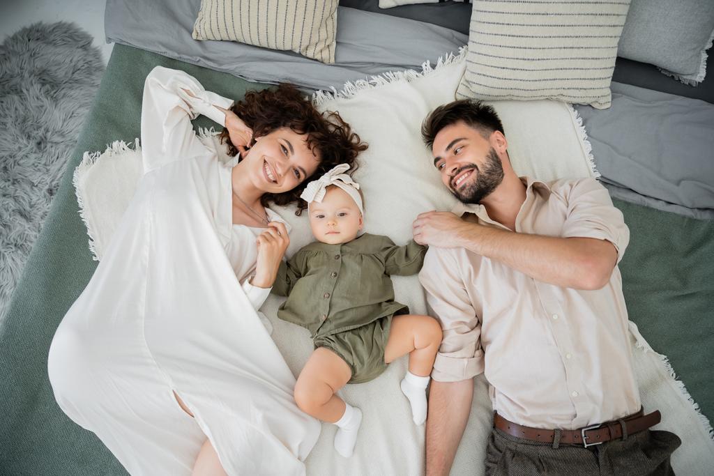 Blick von oben auf freudige Eltern und Baby-Mädchen, die zu Hause auf dem Bett liegen  - Foto, Bild