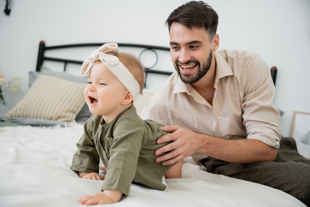 happy bearded father looking at infant baby crawling on bed - Photo, Image