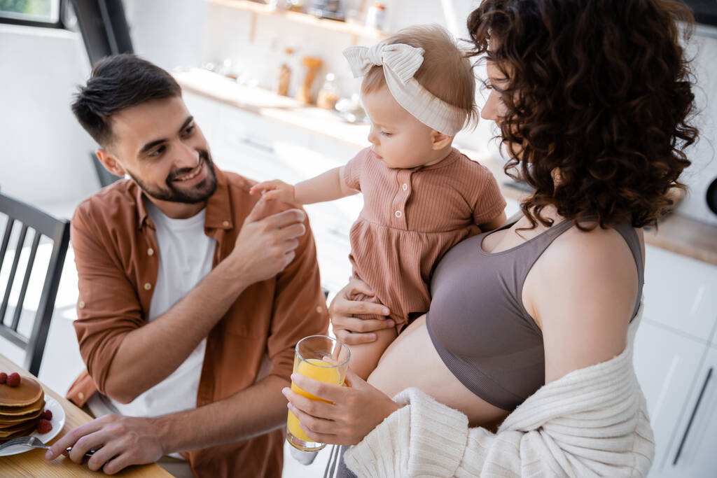 kręcona kobieta trzymająca w ramionach córeczkę i przynosząca mężowi sok pomarańczowy podczas śniadania  - Zdjęcie, obraz