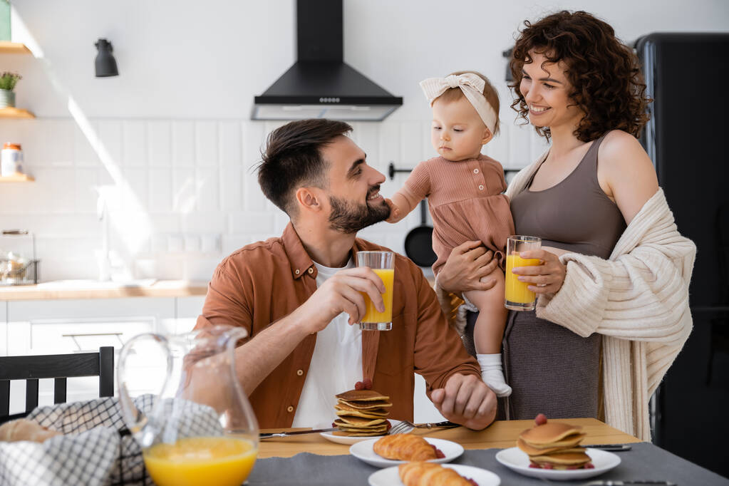 朝食中に赤ちゃんの近くでオレンジジュースを持っている幸せな両親  - 写真・画像