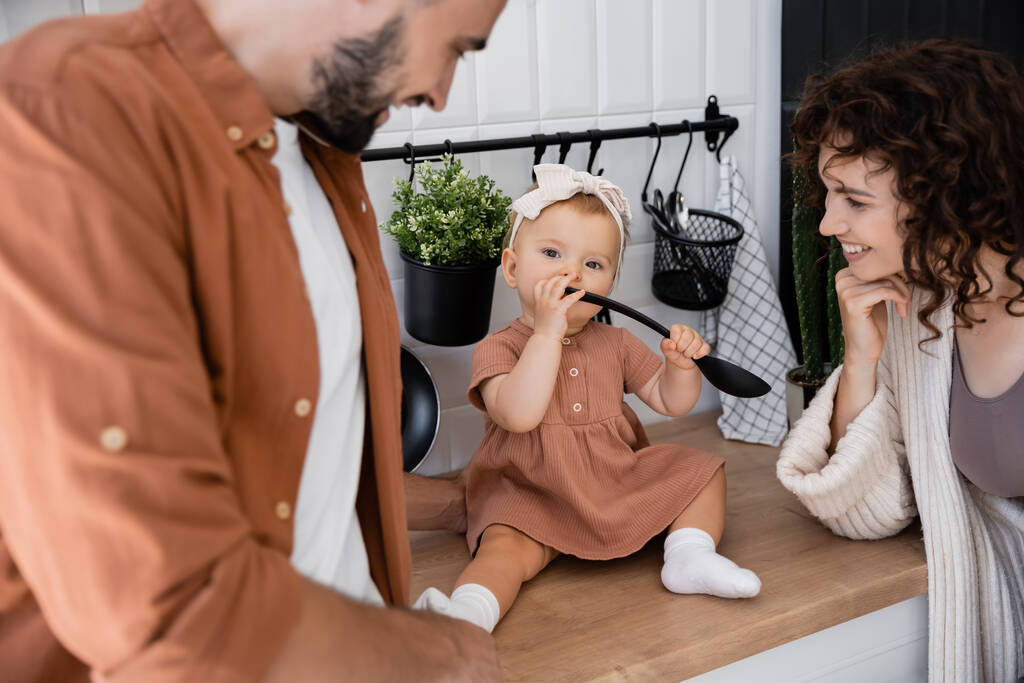 niño pequeño en diadema sosteniendo cucharón cerca de la boca mientras está sentado en encimera de la cocina cerca de padres alegres  - Foto, imagen