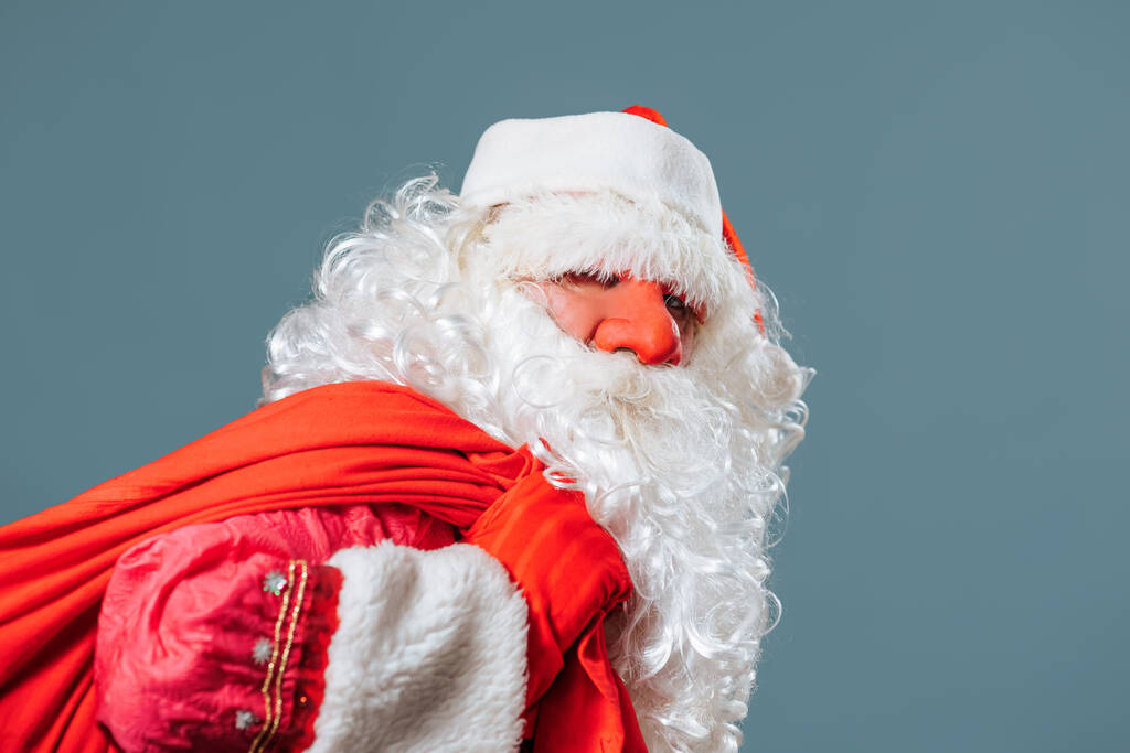 Studio Shooting Seitenansicht blauer Hintergrund müde Weihnachtsmann wirft rote Tasche mit Geschenken auf seine Schultern. Der Weihnachtsmann hält eine schwere Tüte in der Hand. Väterchen Frost im Profil hat bald Geschenke. - Foto, Bild