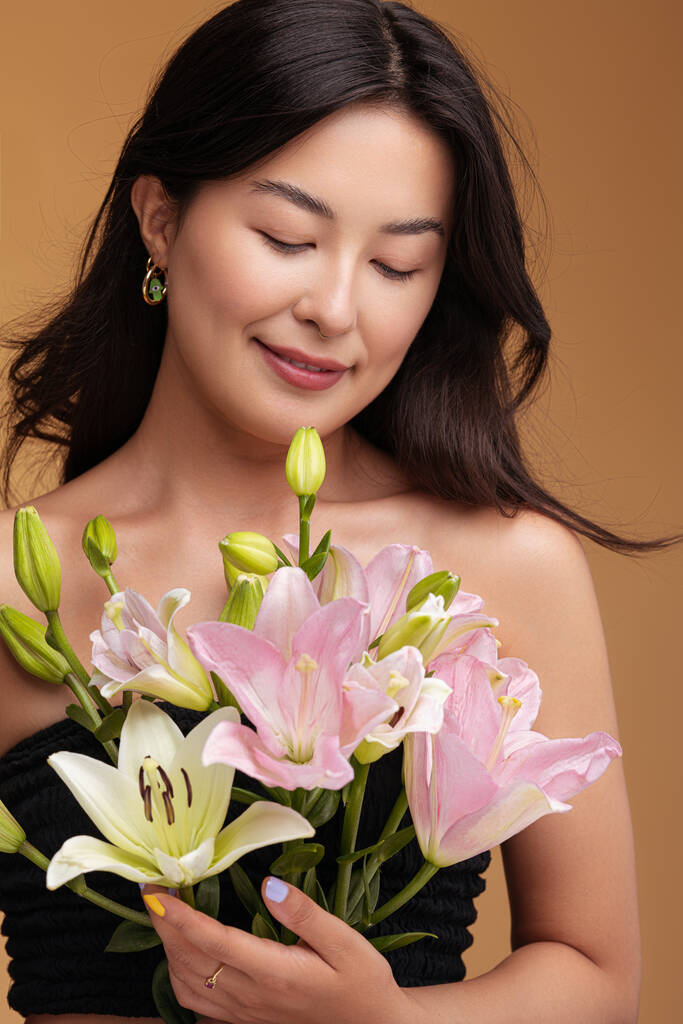 Felice donna asiatica con spalle nude ammirando mazzo di fiori di giglio freschi mentre in piedi contro marrone sfondo marrone - Foto, immagini