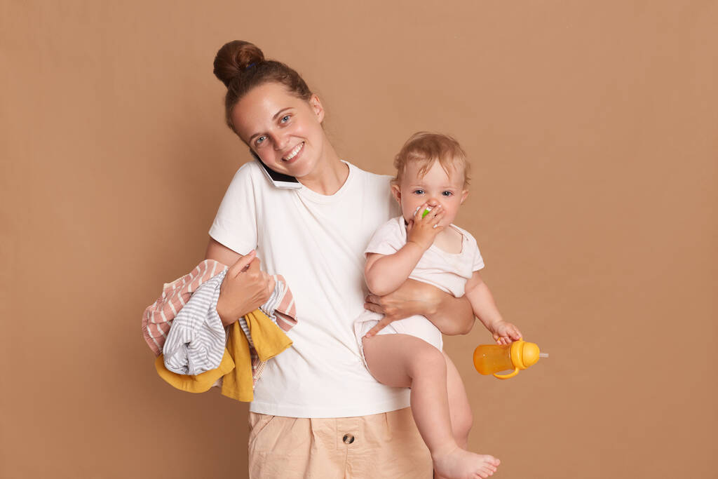 Πορτρέτο της πολυάσχολης μητέρας κρατώντας την κόρη του μωρού και τα ρούχα που απομονώνονται σε καφέ φόντο, θηλυκό φορώντας λευκό t πουκάμισο μιλάμε στο κινητό τηλέφωνο, εκφράζοντας θετικά συναισθήματα. - Φωτογραφία, εικόνα