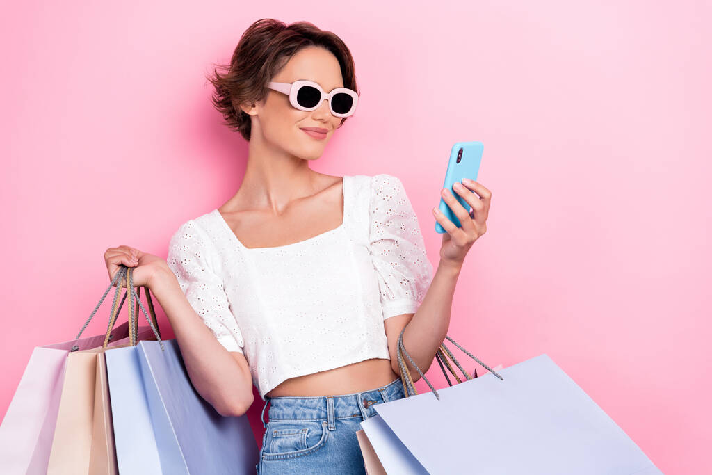 Foto de hermosa chica glamour pelo castaño blanco superior gafas de sol mantener teléfono inteligente compras en línea aislado en el fondo de color rosa. - Foto, imagen