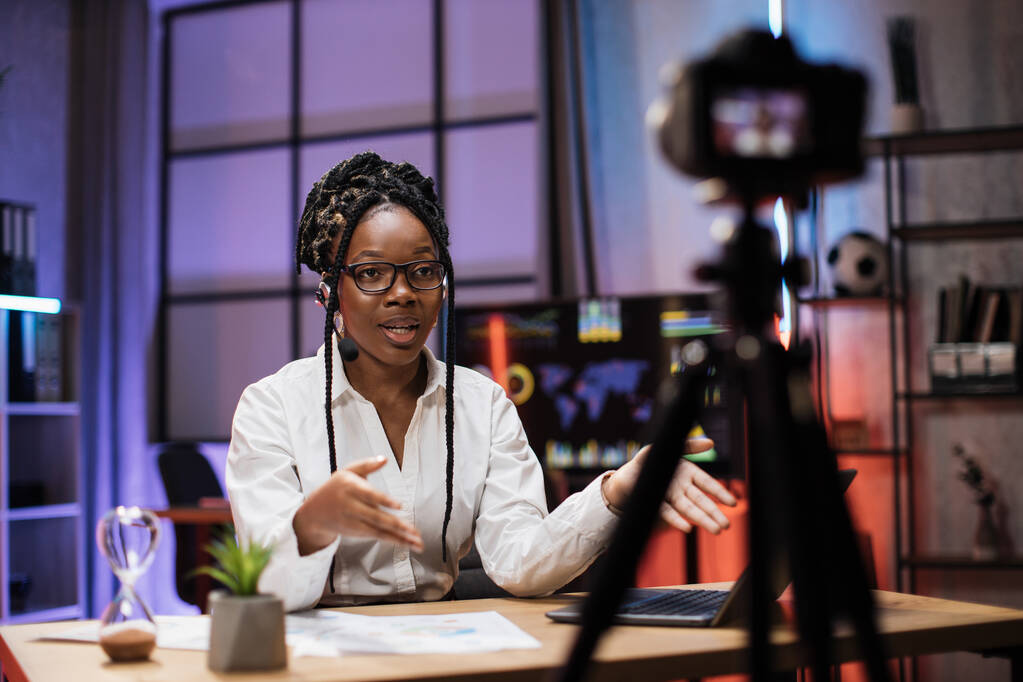 Portret van aantrekkelijke zelfverzekerde ervaren vrouwelijke Afro-Amerikaanse office manager in headset opname video vlog voor internet publiek in avondkantoor. - Foto, afbeelding