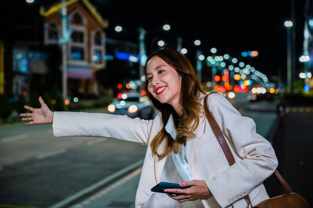 Hermosa mujer sonriendo usando aplicación de teléfono inteligente que llama con la mano hacia arriba taxi después del trabajo tardío, mujer de negocios asiática de pie granizo saludando taxi de la mano en la carretera en la calle de la ciudad ocupada por la noche - Foto, imagen