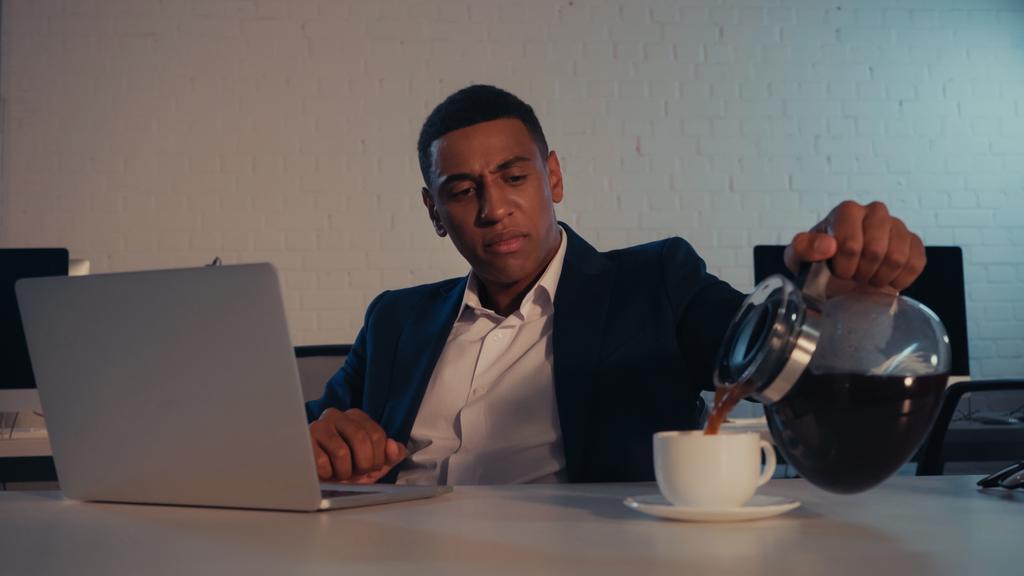 アフリカ系アメリカ人のビジネスマンが夜にオフィスでノートパソコンの近くにコーヒーを注ぐ - 写真・画像
