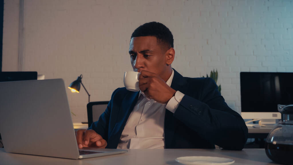 Αφροαμερικάνος επιχειρηματίας πίνοντας καφέ και χρησιμοποιώντας φορητό υπολογιστή στο γραφείο τη νύχτα - Φωτογραφία, εικόνα