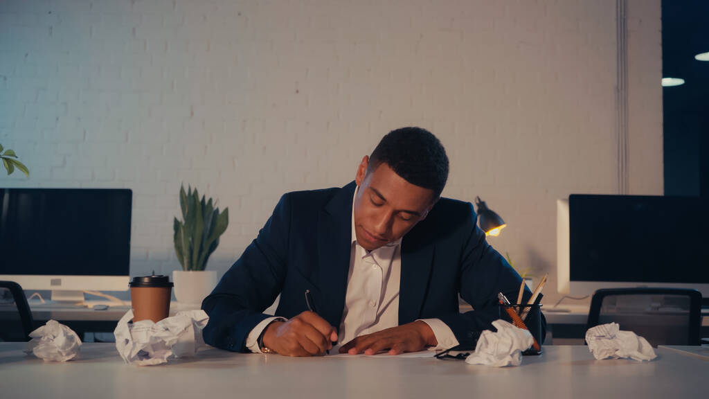 Empresario afroamericano escribiendo en cuaderno cerca de papel arrugado y café para llevar por la noche - Foto, imagen