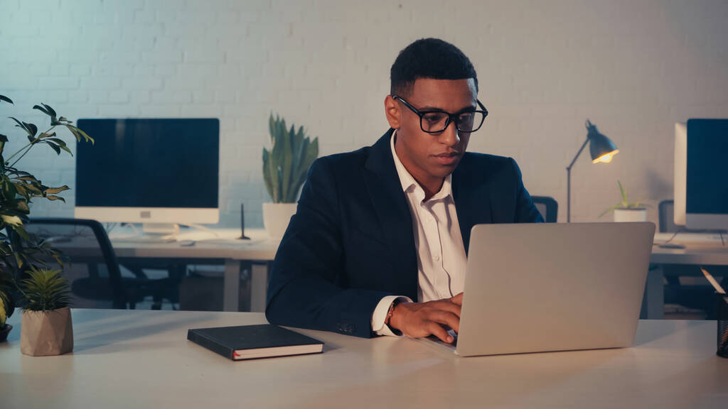 African american manager in eyeglasses using laptop near notebook in office at night - Photo, Image