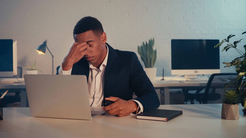 Homme d'affaires afro-américain fatigué assis près d'un ordinateur portable et d'un ordinateur portable au bureau le soir  - Photo, image