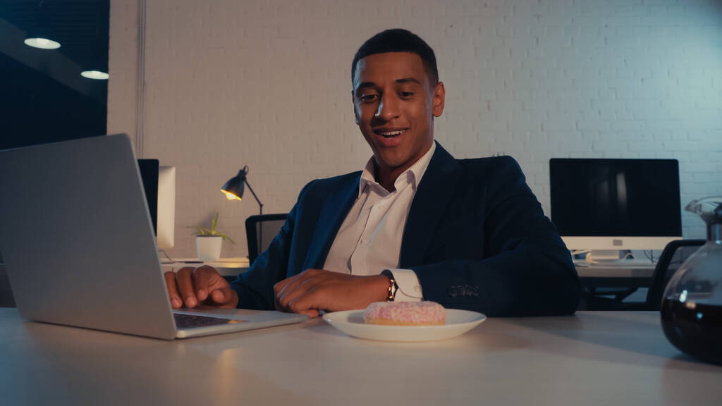 Посмішка афроамериканського бізнесмена, який дивиться на пончик біля кави і ноутбука в офісі.  - Фото, зображення