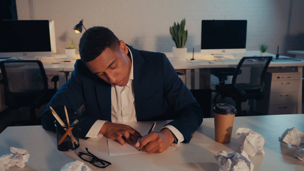 Afrykański amerykański biznesmen piszący na dokumencie przy pogniecionym papierze i kawie w biurze wieczorem  - Zdjęcie, obraz